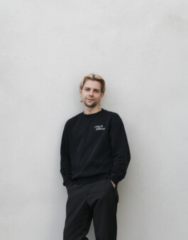 Jakob Eichhorn, a young man in a black outfit stands confidently with his hands in pockets against a plain, light gray wall, smiling subtly. Jakob Eichhorn