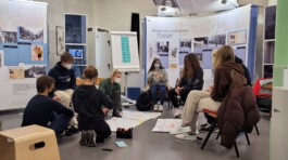 A group of students gathered in a classroom setting, seated on chairs and the floor, attentively engaged in a discussion surrounded by educational displays and posters. Jakob Eichhorn - Cultural Communication
